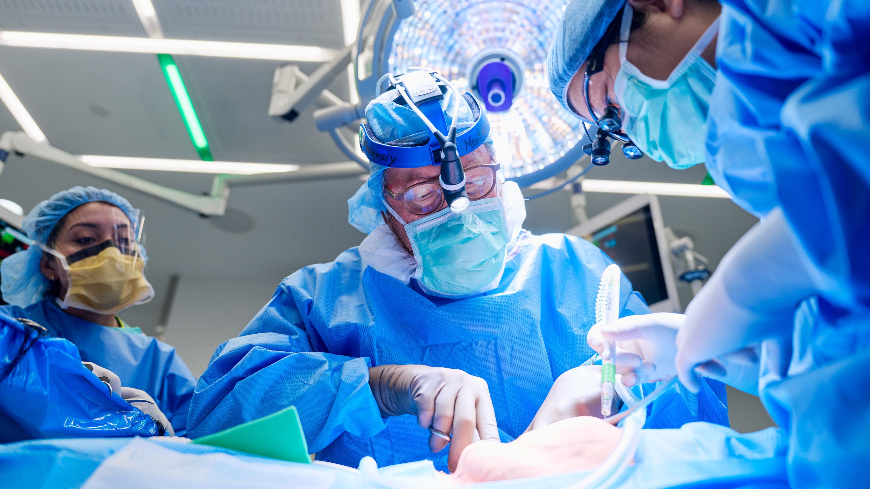 A surgeon attended by two other medical professionals prepares the patient draped in the foreground